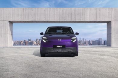 Photograph showcasing a purple Nissan Pixo concept car parked before a concrete architectural opening with a city skyline beyond. Front view highlights illuminated angular headlights, closed grille, PIXO plate, sleek compact design, and blue sky backdrop.